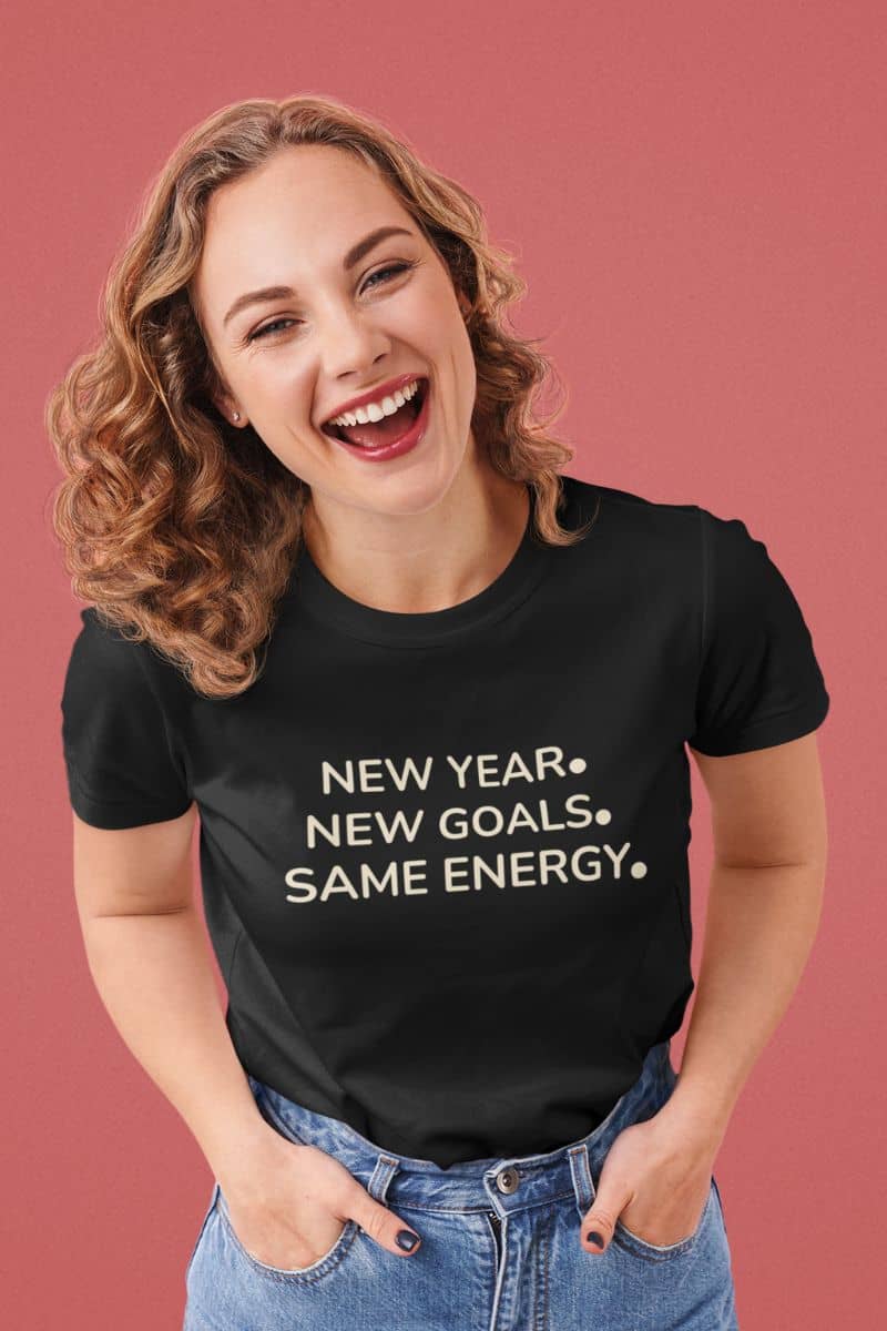 Funny new year shirt with a woman wearing a black t-shirt with 'NEW YEAR. NEW GOALS. SAME ENERGY.' text on a pink background
