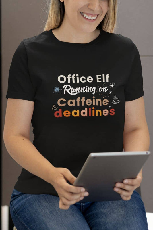 Funny office shirt with a person wearing a black t-shirt with colorful text 'Office Elf, Running on Caffeine & Deadlines' holding a tablet.