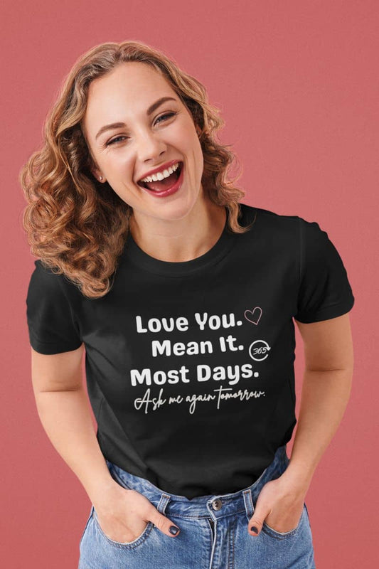 Funny relationship shirt with a woman wearing a black t-shirt with text 'Love You. Mean It. Most Days. Ask me again tomorrow.' on a pink background
