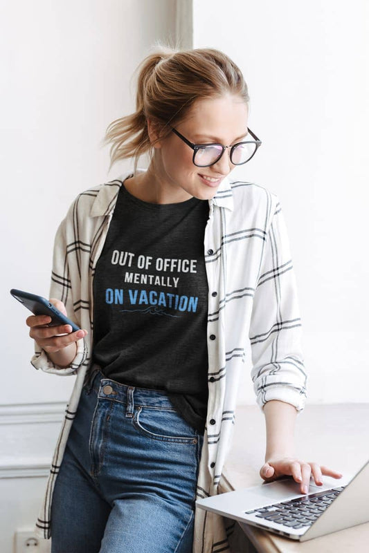 Funny vacation t-shirt with a woman wearing a black t-shirt with text 'Out of Office: Mentally on Vacation', using a laptop and phone indoors.