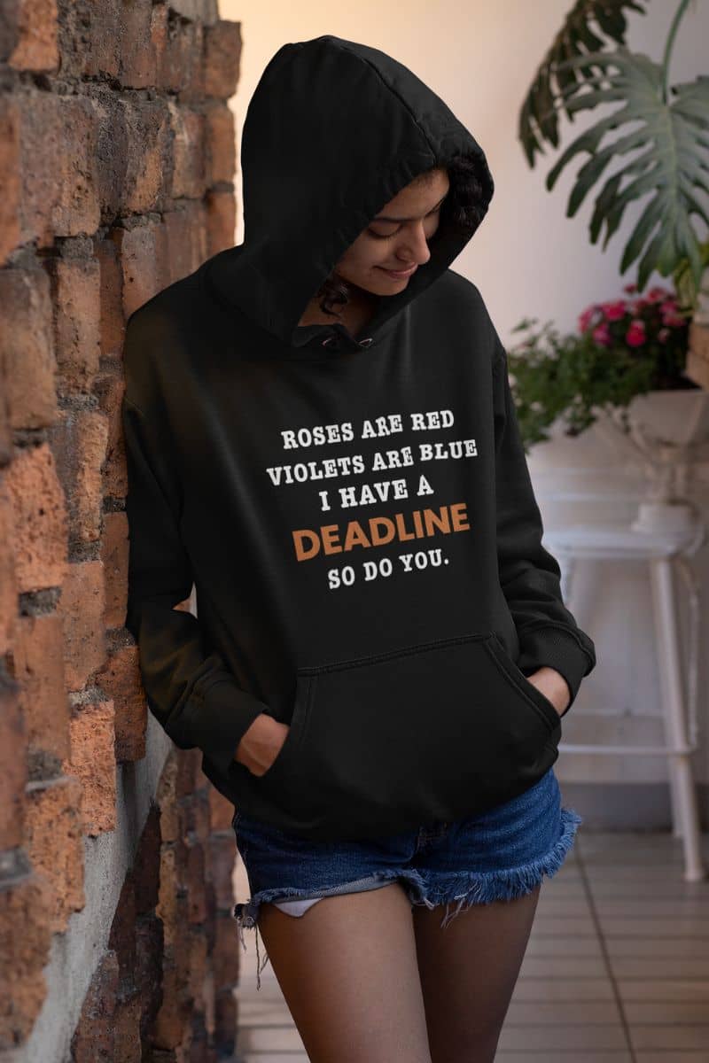Funny work hoodie with a person wearing a black hoodie with text 'Roes are red, violets are blue. I have a deadline so you do' leaning against a brick wall.