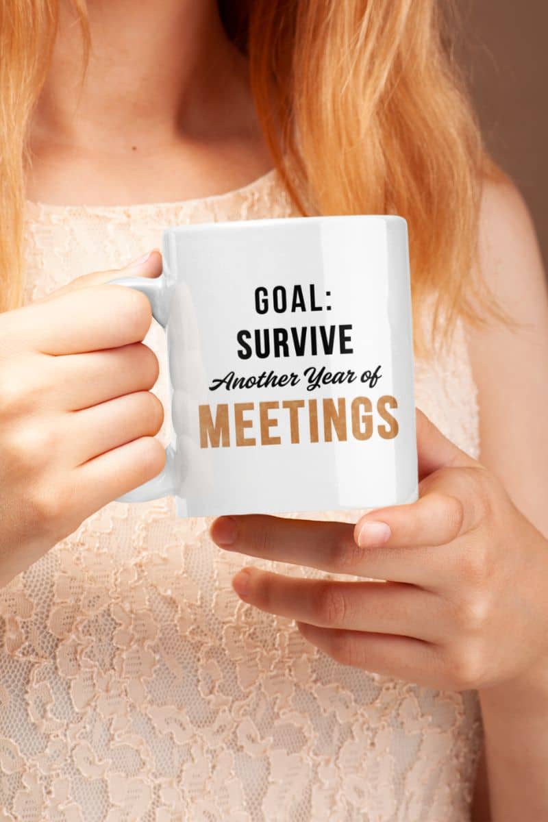 Funny work mugs with a person holding a mug with text 'Goal: Survive Another Year of Meetings' against a neutral background