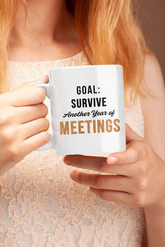 Funny work mugs with a person holding a mug with text 'Goal: Survive Another Year of Meetings' against a neutral background