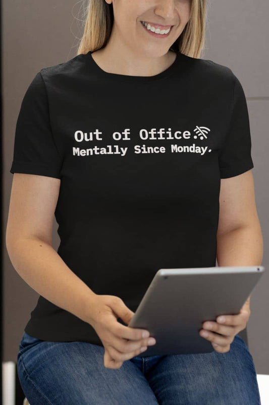 Funny work quote t-shirt with a person wearing a black t-shirt with text 'Out of Office. Mentally Since Monday.' holding a tablet.