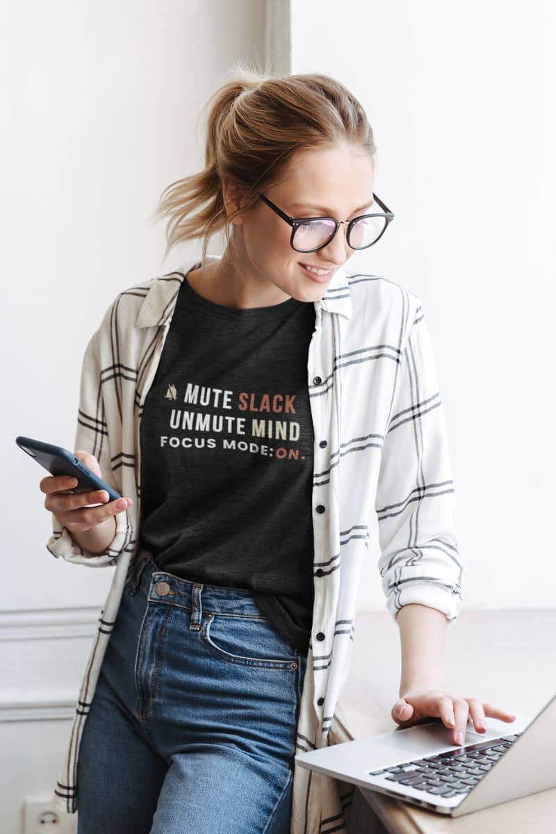 Funny work t shirt for mental health with a woman wearing a black t-shirt with text 'Mute Slack, Unmute Mind. Focus Mode: On', using a laptop and phone indoors.