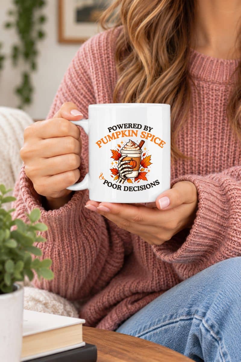 A woman holding a Halloween coffee mug with the text 'Powered by Pumpkin Spice & Poor Decisions', sitting at the desk