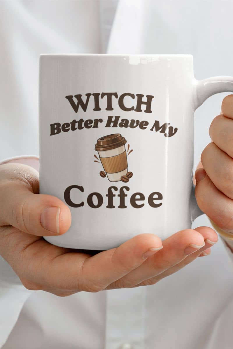 Halloween coffee mug with a white ceramic coffee mug featuring the text 'Witch Better Have My Coffee' and a coffee cup graphic, held in two hands against a white shirt background.
