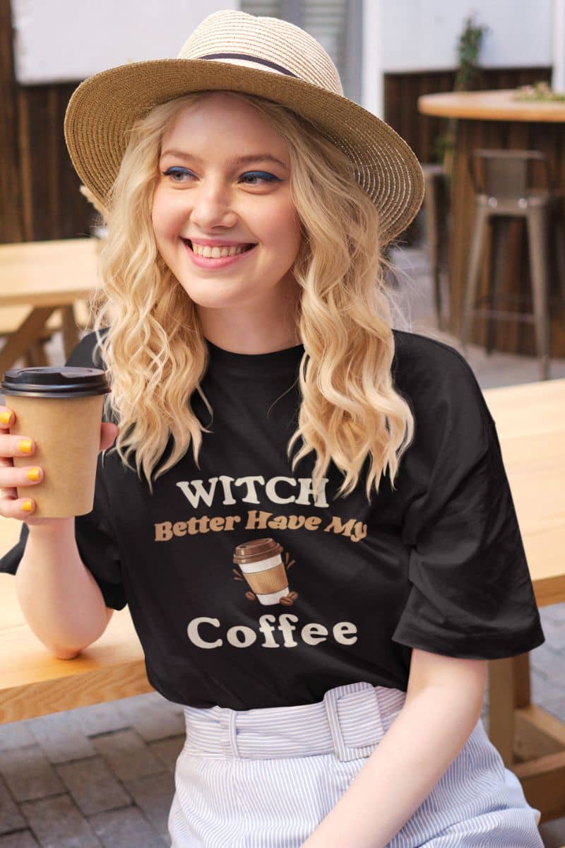 Halloween coffee shirt with a  woman wearing a straw hat and a black graphic T-shirt that reads 'Witch Better Have My Coffee', holding a takeaway coffee cup in an outdoor cafe setting.