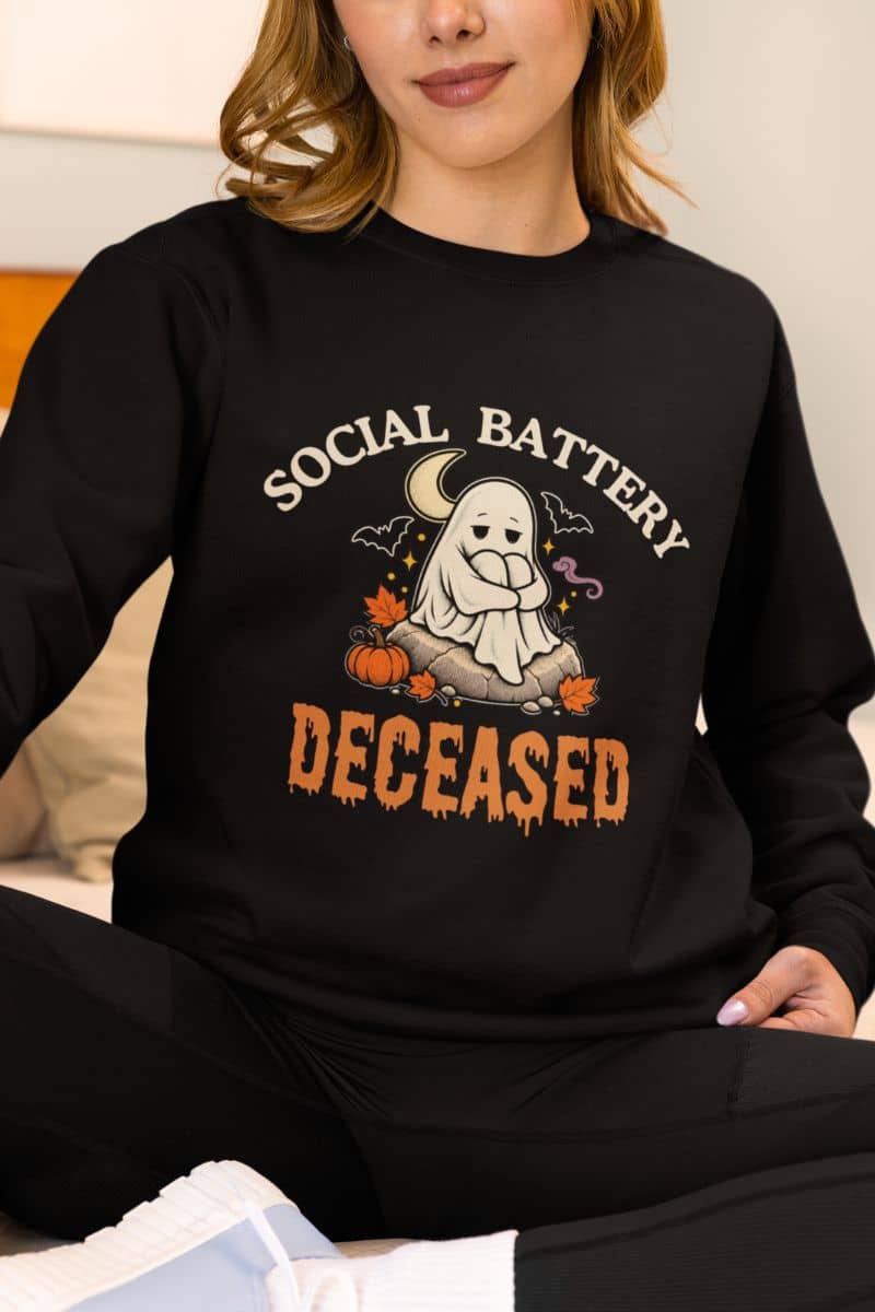A woman wearing a Halloween crewneck featuring a 'Social Battery: Deceased' graphic with a ghost sitting around decorative Halloween items