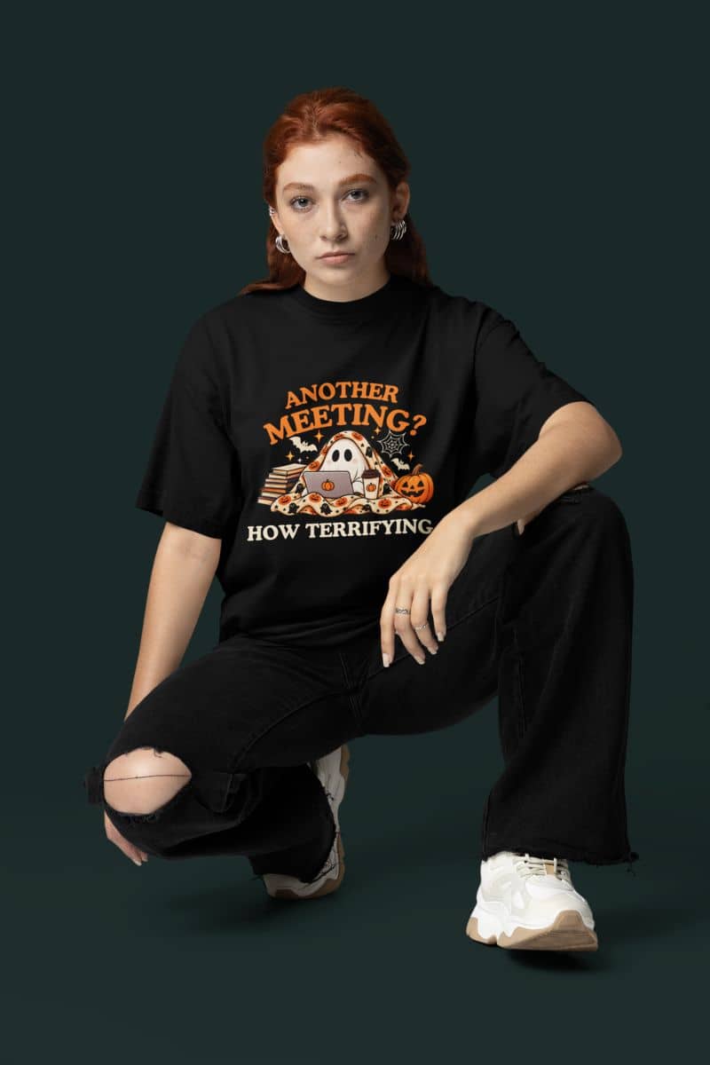 A model with red hair crouches in a studio setting wearing a Halloween ghost with a Halloween-themed design that reads 'Another Meeting? How Terrifying.'