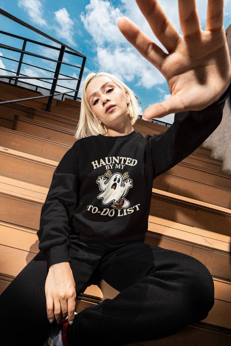 A woman wearing a Halloween ghost sweatshirt featuring a “Haunted by My To-Do List” funny ghost, sitting on the steps