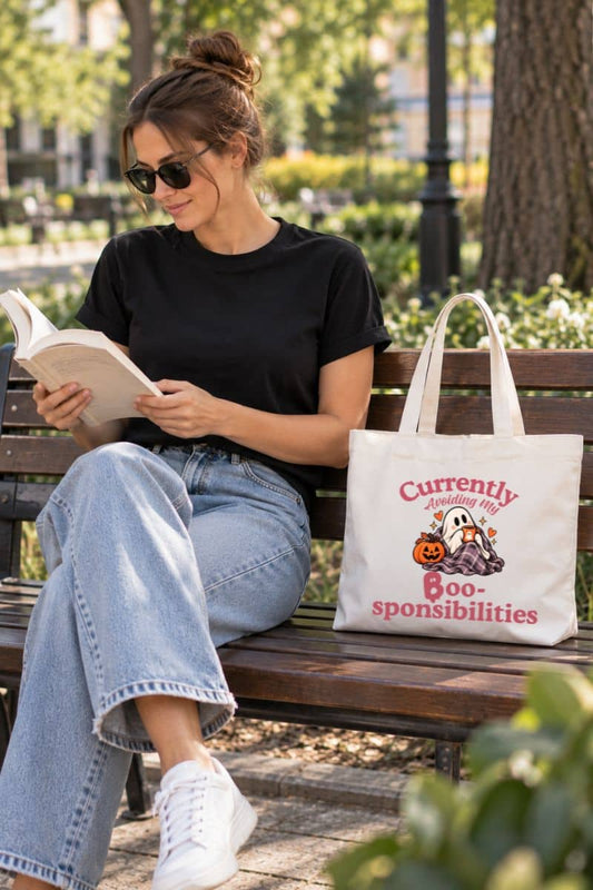 A woman in a black t-shirt and jeans sits on a park bench reading a book, with a Halloween ghost tote bag featuring a Halloween graphic and the text 'Currently Avoiding My Boo-sponsibilities' beside her.