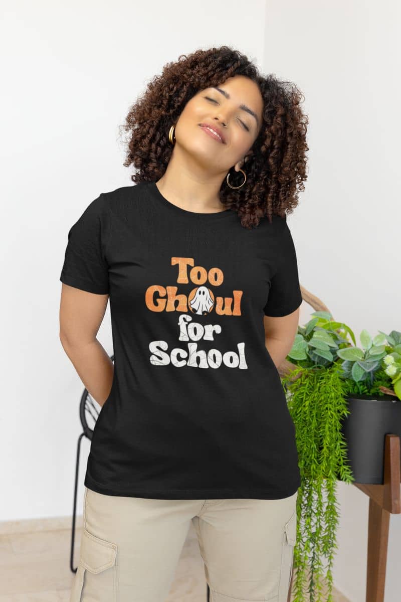 Halloween graphic tee for women with a person in a black t-shirt with 'Too Ghoul for School' text, standing indoors with plants in the background.
