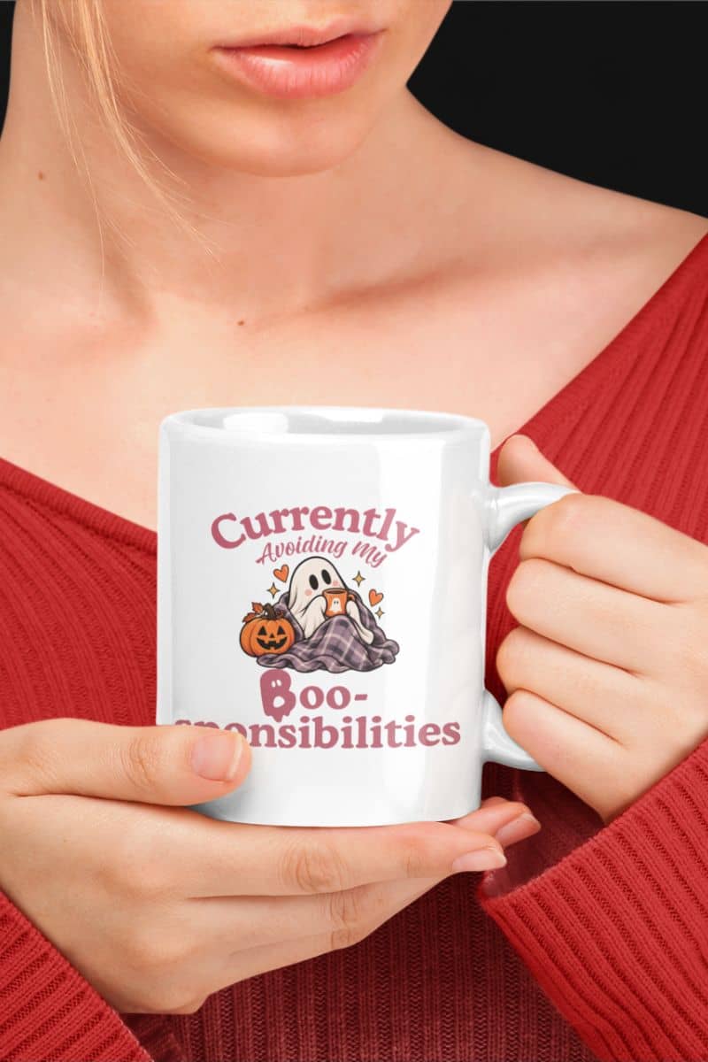 A person in a red ribbed sweater holding a Halloween mug with a Halloween graphic that reads 'Currently Avoiding My Boo-sponsibilities' with a ghost and pumpkin.