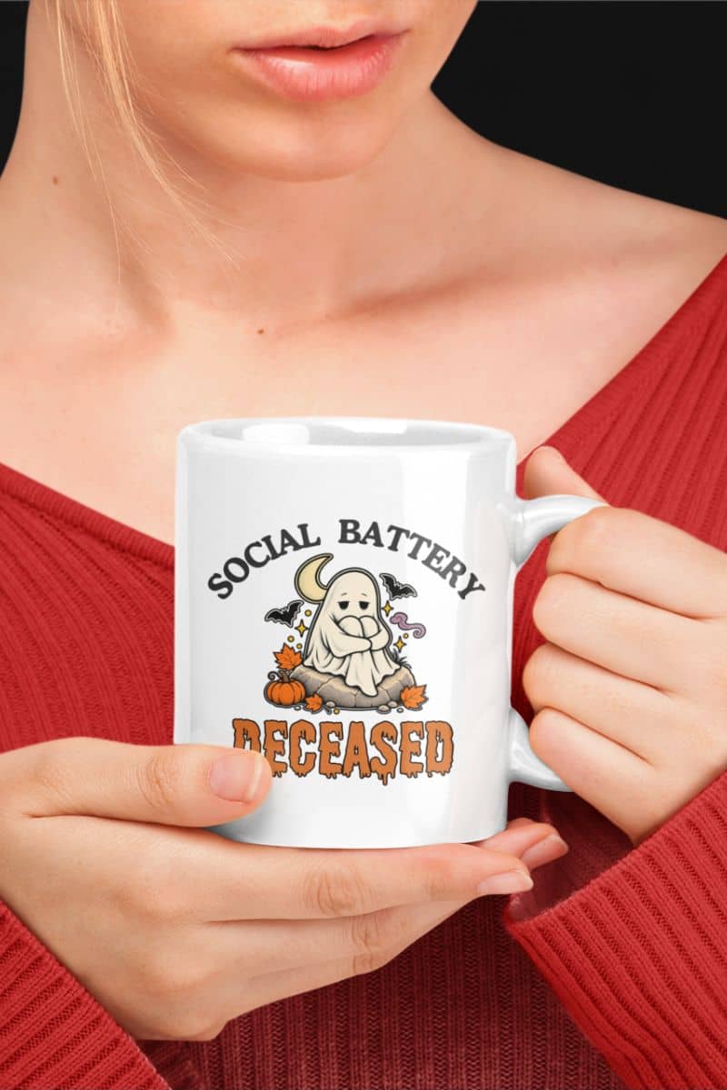 A person in a red ribbed sweater holding a Halloween office mug featuring a 'Social Battery: Deceased' graphic with a  ghost and pumpkins