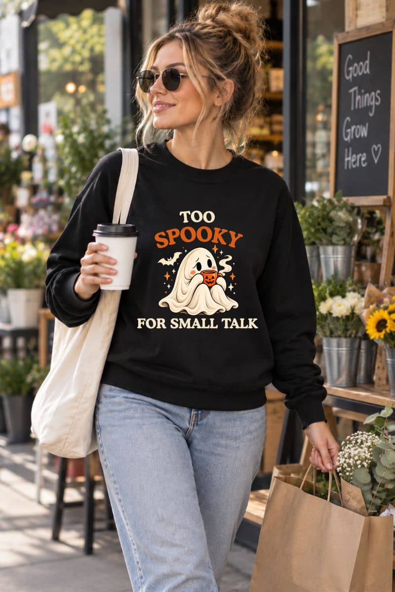 Woman wearing a black Halloween Pullover with the “Too Spooky for Small Talk” ghost design while carrying coffee and shopping bags outdoors.
