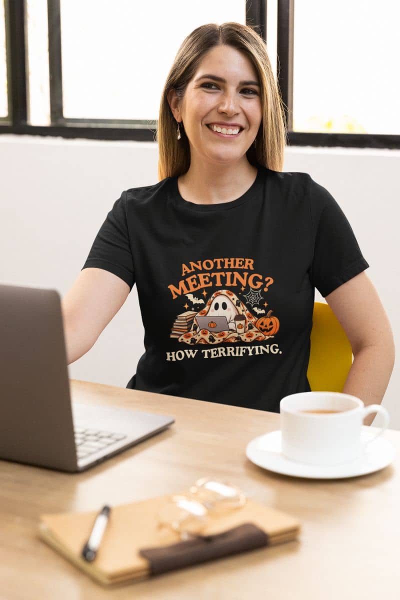 A woman smiling at a desk wearing a Halloween shirt with a Halloween-themed design that reads 'Another Meeting? How Terrifying.'