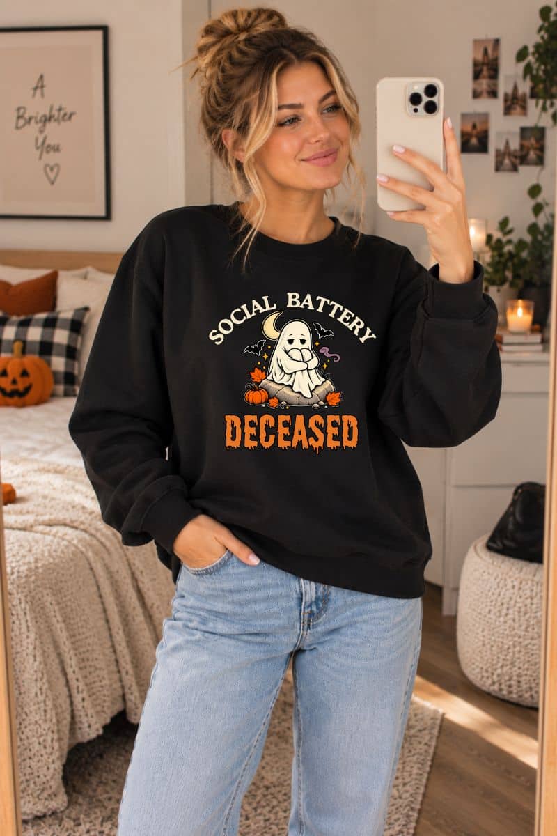 A Halloween sweatshirt for adults worn by a woman, featuring the text 'Social Battery: Deceased' and a ghost sitting around decorative Halloween items
