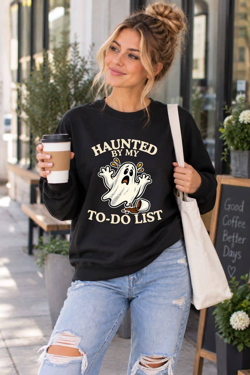 A woman standing in front of a coffee shop, wearing a Halloween sweatshirt featuring a “Haunted by My To-Do List” funny ghost 