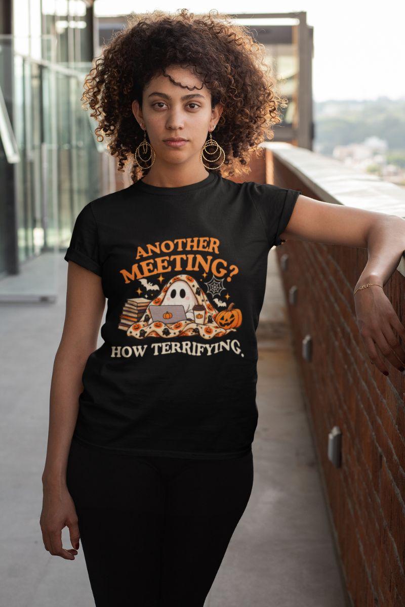 A woman wearing a Halloween tee featuring a 'Another Meeting? How Terrifying.' graphic with a ghost and cup of coffee, standing in the hallway