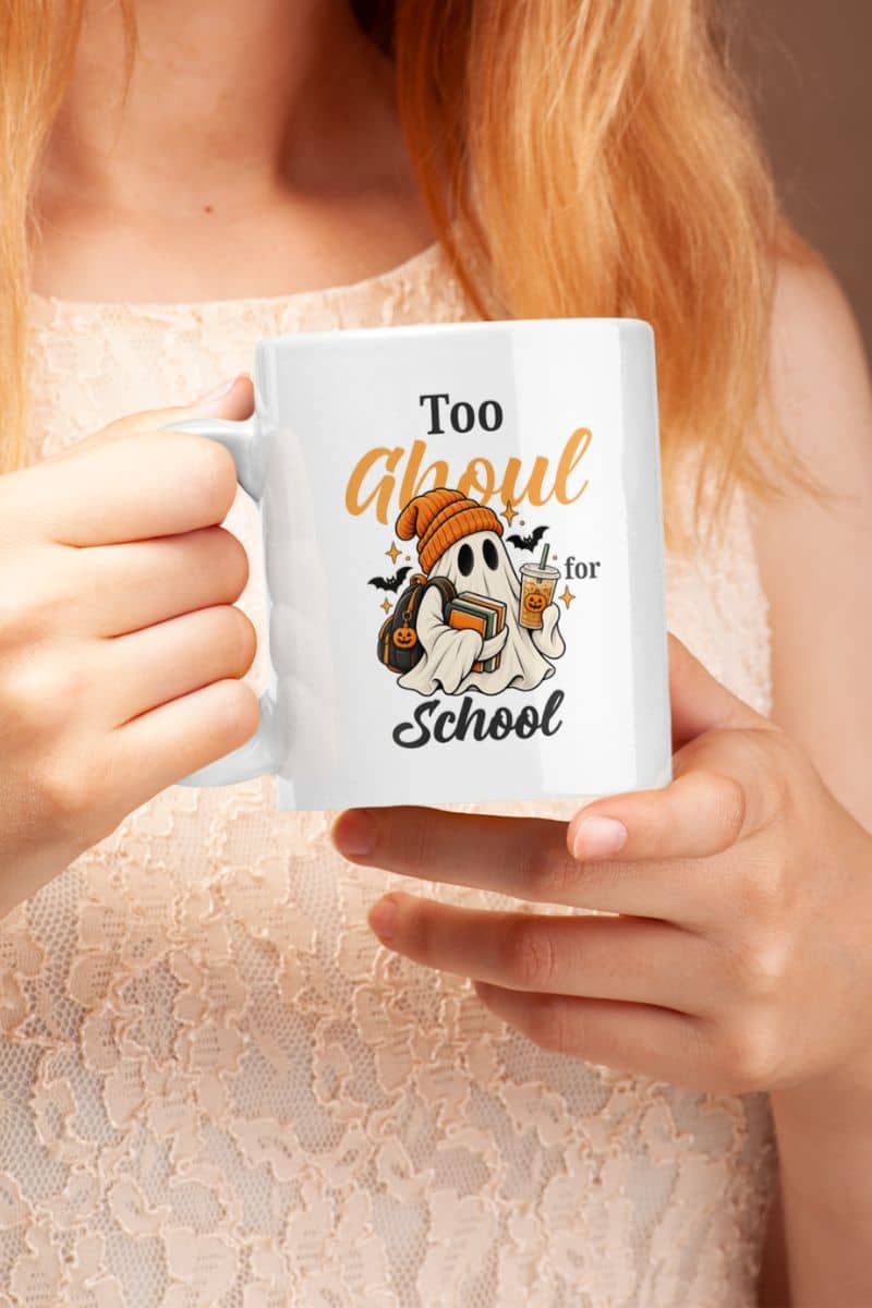 A person holding a white Halloween themed mug that reads 'Too Ghoul for School' featuring a ghost holding books and coffee, wearing a light beige lace top.