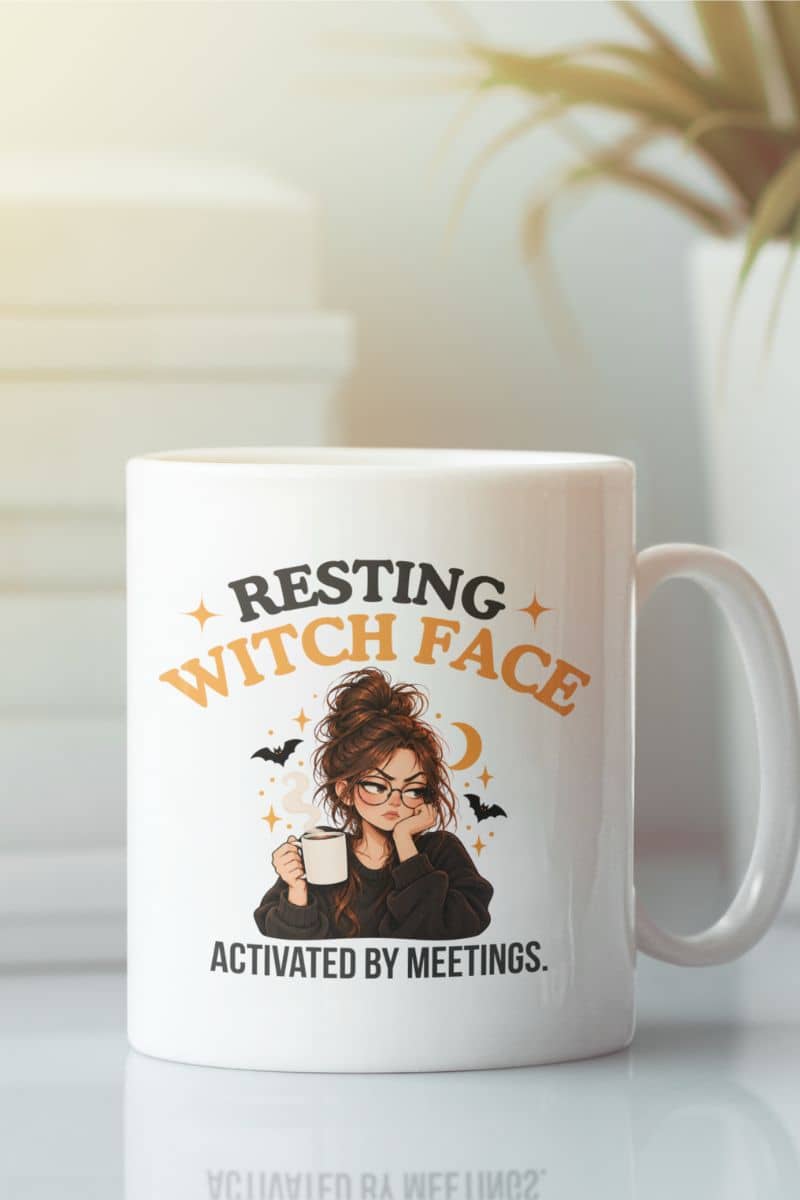 A white Halloween themed mug with a humorous graphic that reads 'Resting Witch Face. Activated by Meetings.' featuring an illustration of a witch holding a mug, surrounded by stars, bats, and a crescent moon, set against a softly blurred background.