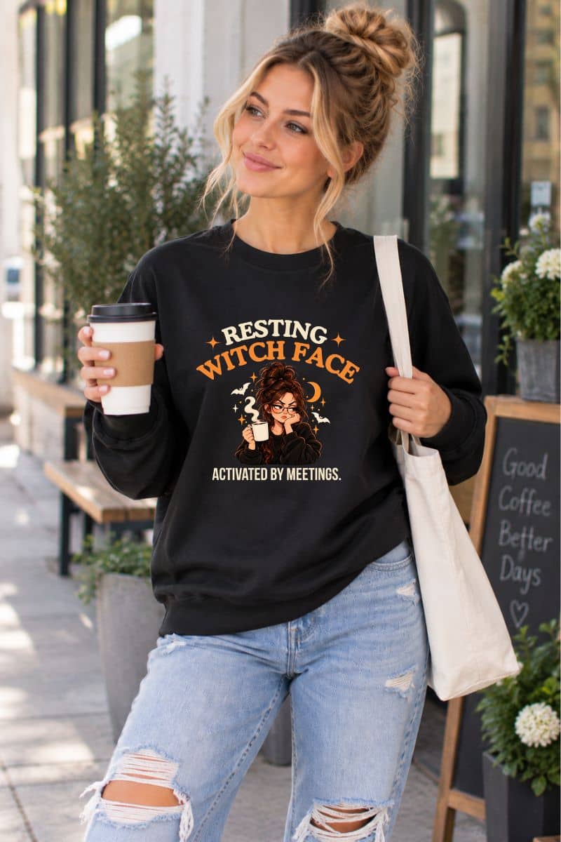 A woman standing in front of a coffee shop wearing a sweatshirt featuring the text 'Resting Witch Face. Activated by Meetings.' with a woman's face