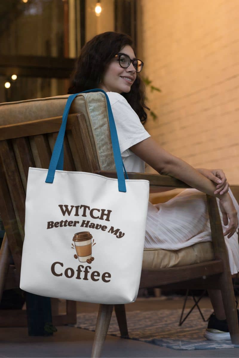 A Halloween tote bag with blue handles and a brown “Witch Better Have My Coffee” graphic, hanging on a wooden chair next to a woman in a cozy cafe setting.