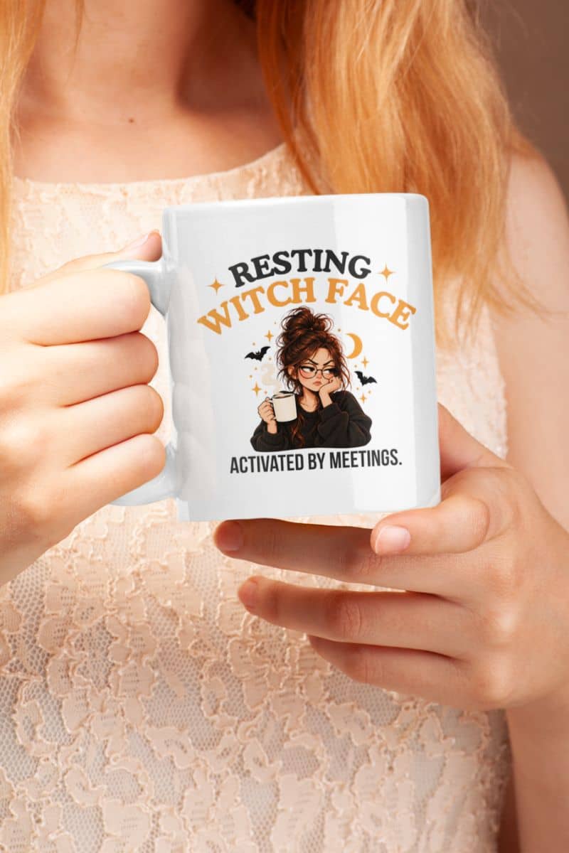 A person holding a Halloween witch mug with a Halloween-themed graphic that reads 'Resting Witch Face. Activated by Meetings.' The person is wearing a light beige lace top, and the mug features an illustration of a witch with glasses and bats.