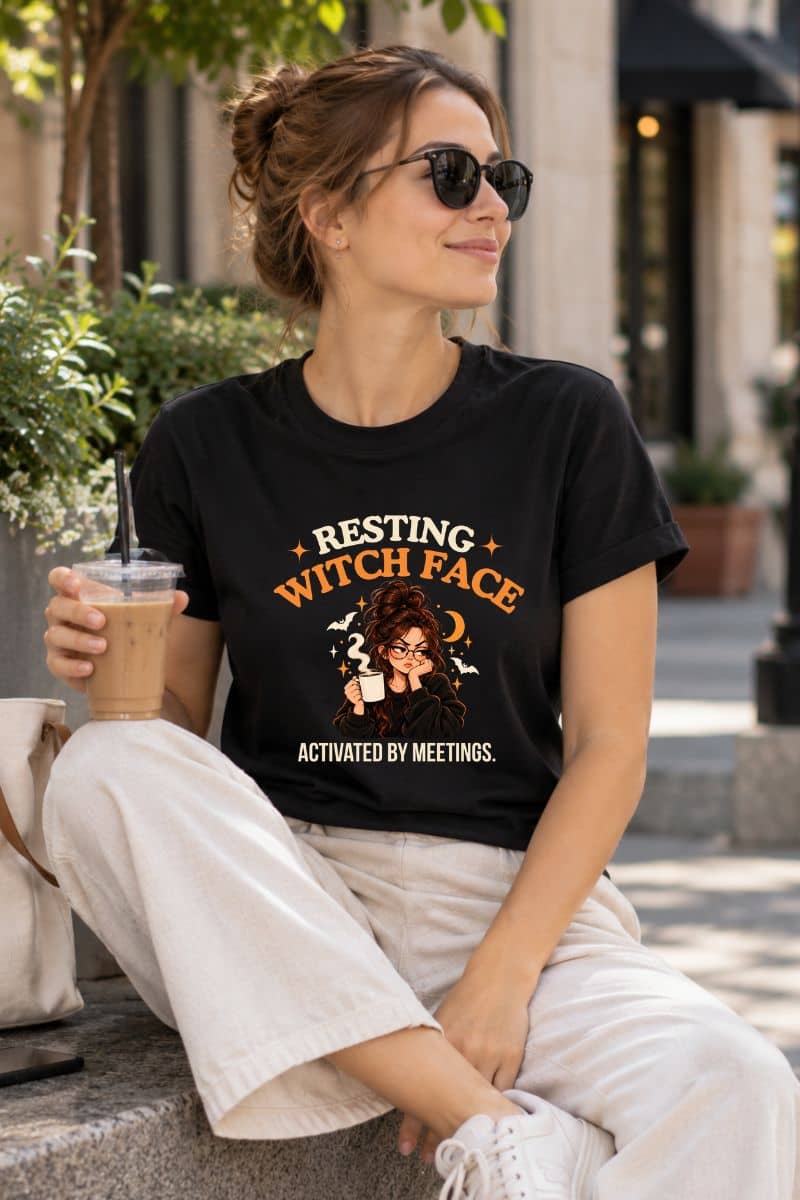 A woman sitting outdoors wearing a Halloween witch shirt featuring a “Resting Witch Face. Activated by Meetings.” graphic with a woman’s face.