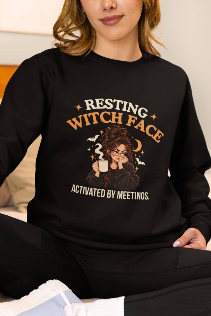 A woman wearing a Halloween witch sweatshirt featuring the text 'Resting Witch Face. Activated by Meetings.' with a woman's face