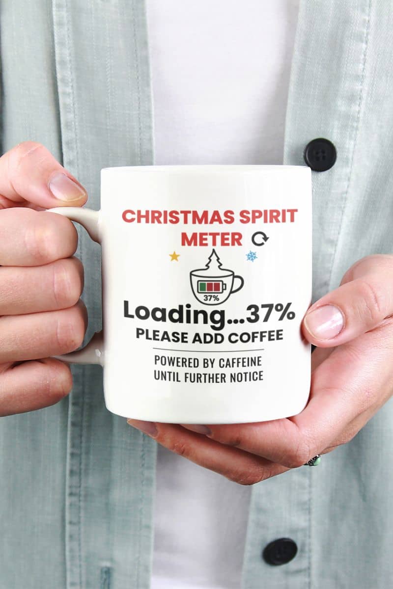 Holiday mug for coworkers with a person holding a mug text 'Christmas Spirit Meter, Loading…37%. Please add coffee. Powered by caffeine until further notice'