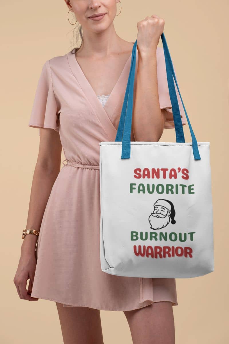 Holiday errand tote bag with a woman holding a tote bag with 'Santa's Favorite Burnout Warrior' text and graphic on a beige background