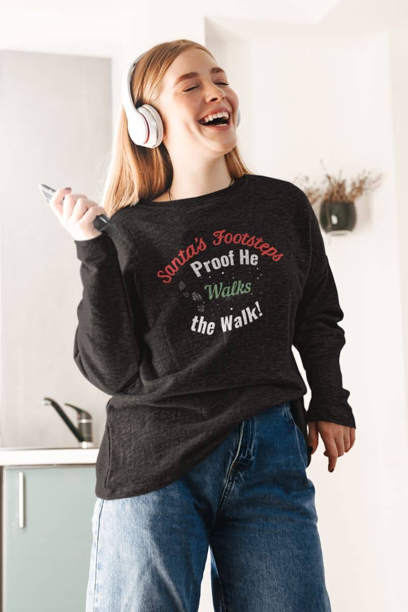 Holiday humor pullover with a person wearing a black sweatshirt with text 'Santa's Footsteps - Proof He Walks The Walk!', standing in a kitchen.