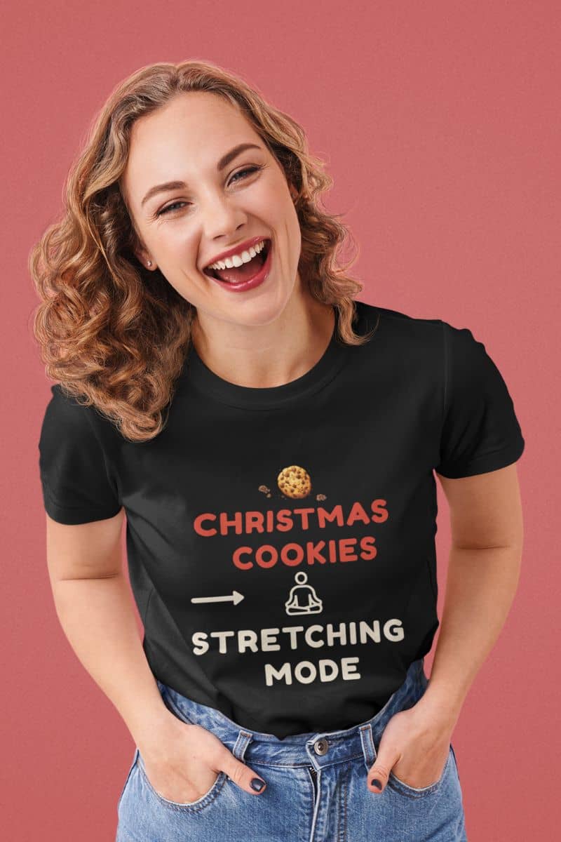 Holiday wellness humor shirt with a woman wearing a black t-shirt with text 'Christmas Cookies, Stretching Mode' on a pink background