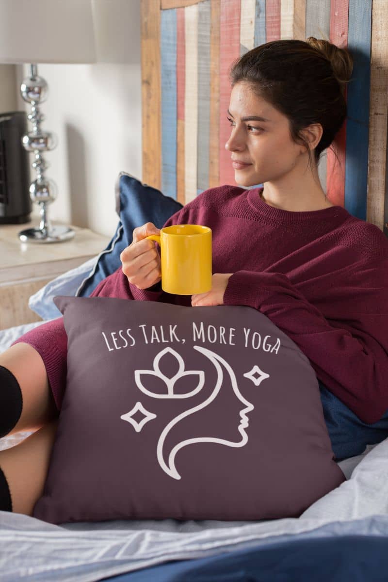 Experience Serenity with the Less Talk More Yoga Pillow with the girl hugging a pillow, holding a cup of tea, pensively on the bed
