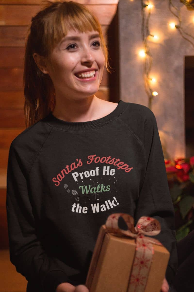 Matching family holiday sweatshirt with a person wearing a black sweater with text 'Santa's Footsteps - Proof He Walks The Walk!', holding a gift box indoors.