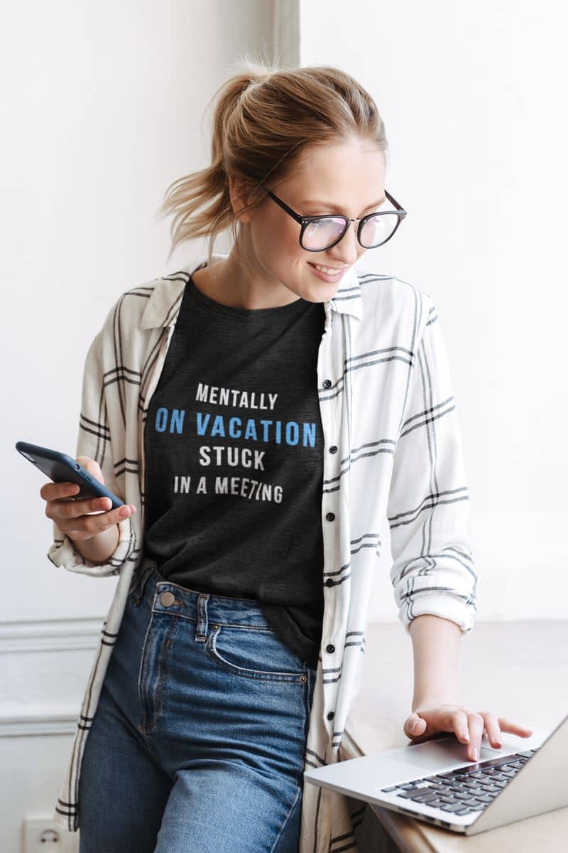 Meeting humor shirt featuring a woman wearing a black t-shirt with text 'Mentally on Vacation. Stuck in A Meeting', standing in an indoor setting.