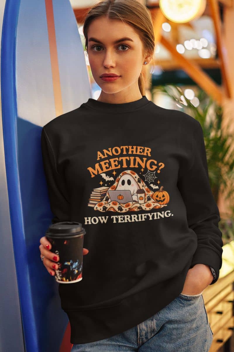 A woman wearing a meeting sweatshirt featuring the text 'Another Meeting? How Terrifying.', holding a cup of coffee