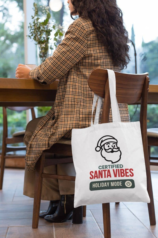 Minimal Christmas tote bag with a person sitting at a table with a tote bag featuring 'Certified Santa Vibes – Holiday Mode On' text.