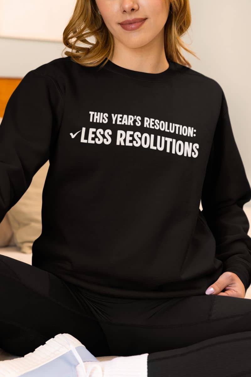 Minimalist humor sweatshirt with a person wearing a black sweatshirt with humorous text 'This year’s resolution: Less resolutions.'