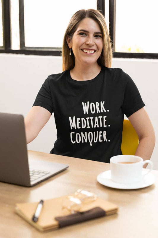 Minimalist motivation t-shirt with a woman wearing a black t-shirt with 'Work. Meditate. Conquer.' text, sitting at a desk with a laptop and coffee.