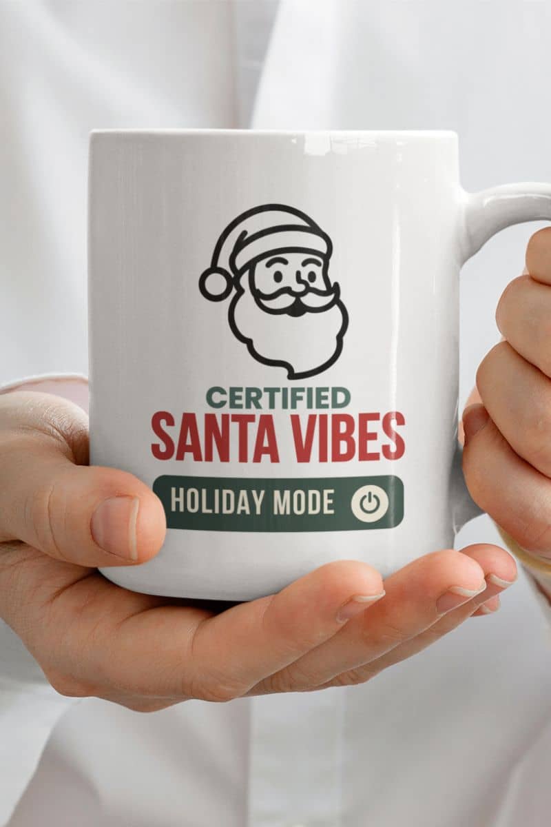 Modern Christmas coffee mug with a mug with Santa Claus design and text 'Certified Santa Vibes Holiday Mode On' held by a person.