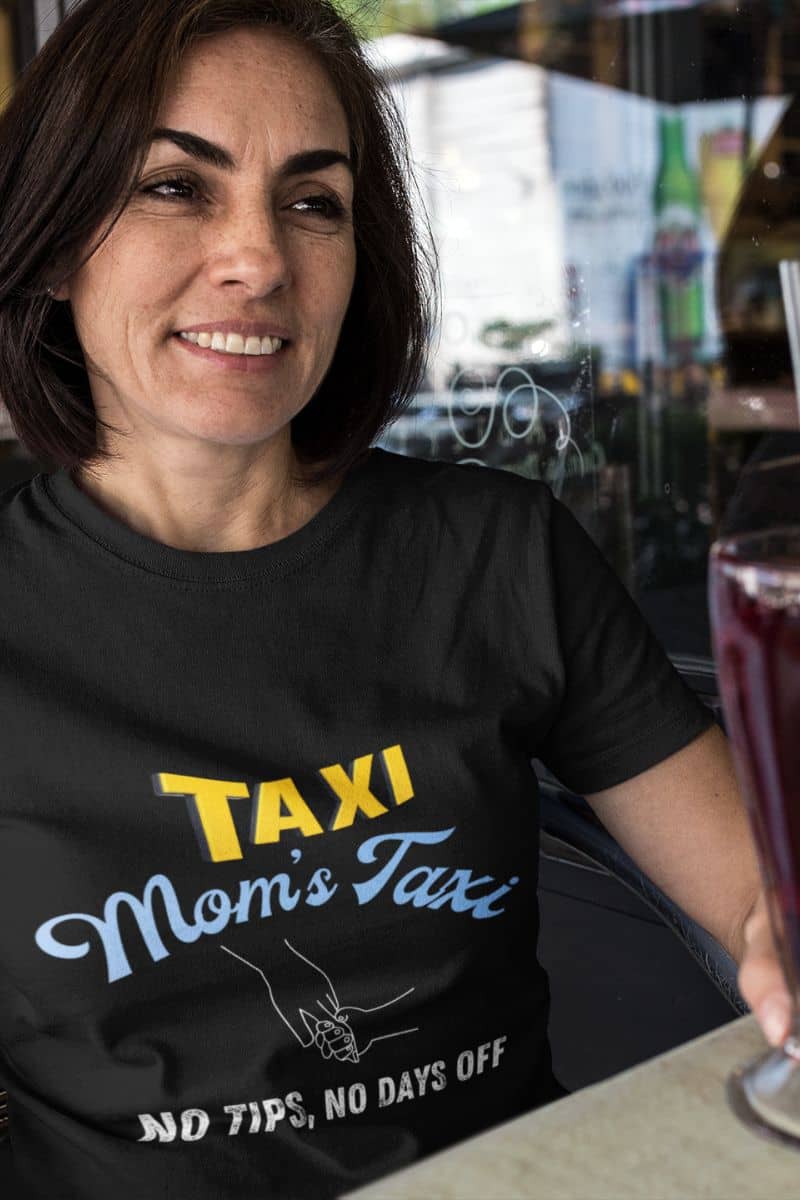 Mom's taxi shirt with a middle aged woman smiling brightly at a cafe with a glass of juice in one hand on the table