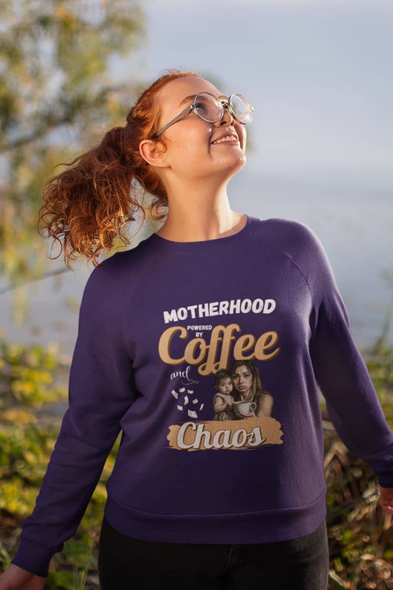 Motherhood quote sweatshirt with a person wearing a purple sweatshirt with text 'Motherhood powered by Coffee and Chaos' and a graphic, standing outdoors.