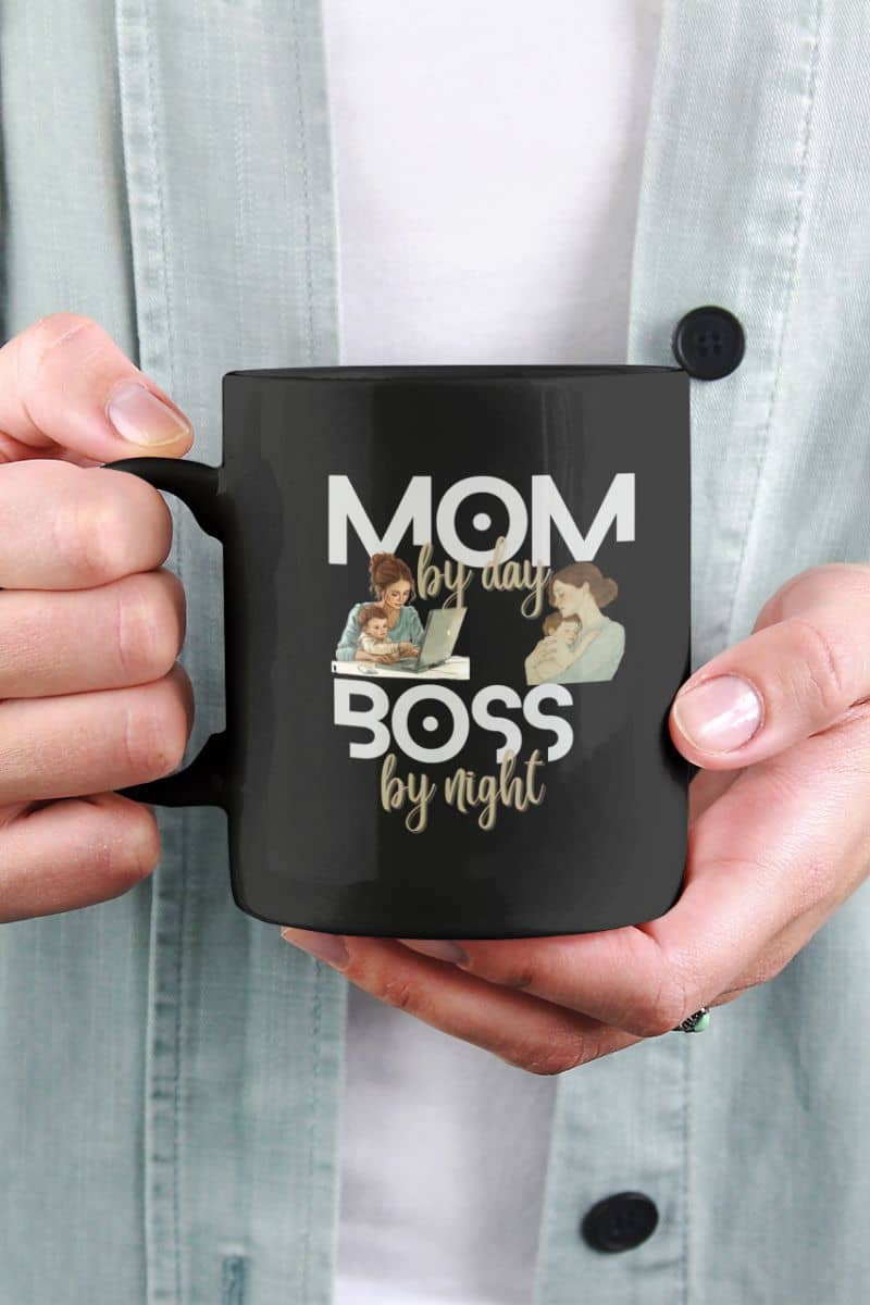 Mother's day coffee mug with a black mug with 'Mom by day, Boss by night' text and images of a mother and child holding a laptop, held by hands against a light gray background.