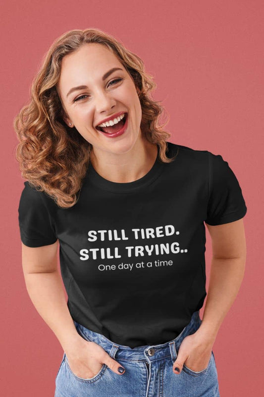 Motivational mental health t-shirt with a woman wearing a black t-shirt with 'STILL TIRED. STILL TRYING. One day at a time.' text on a pink background
