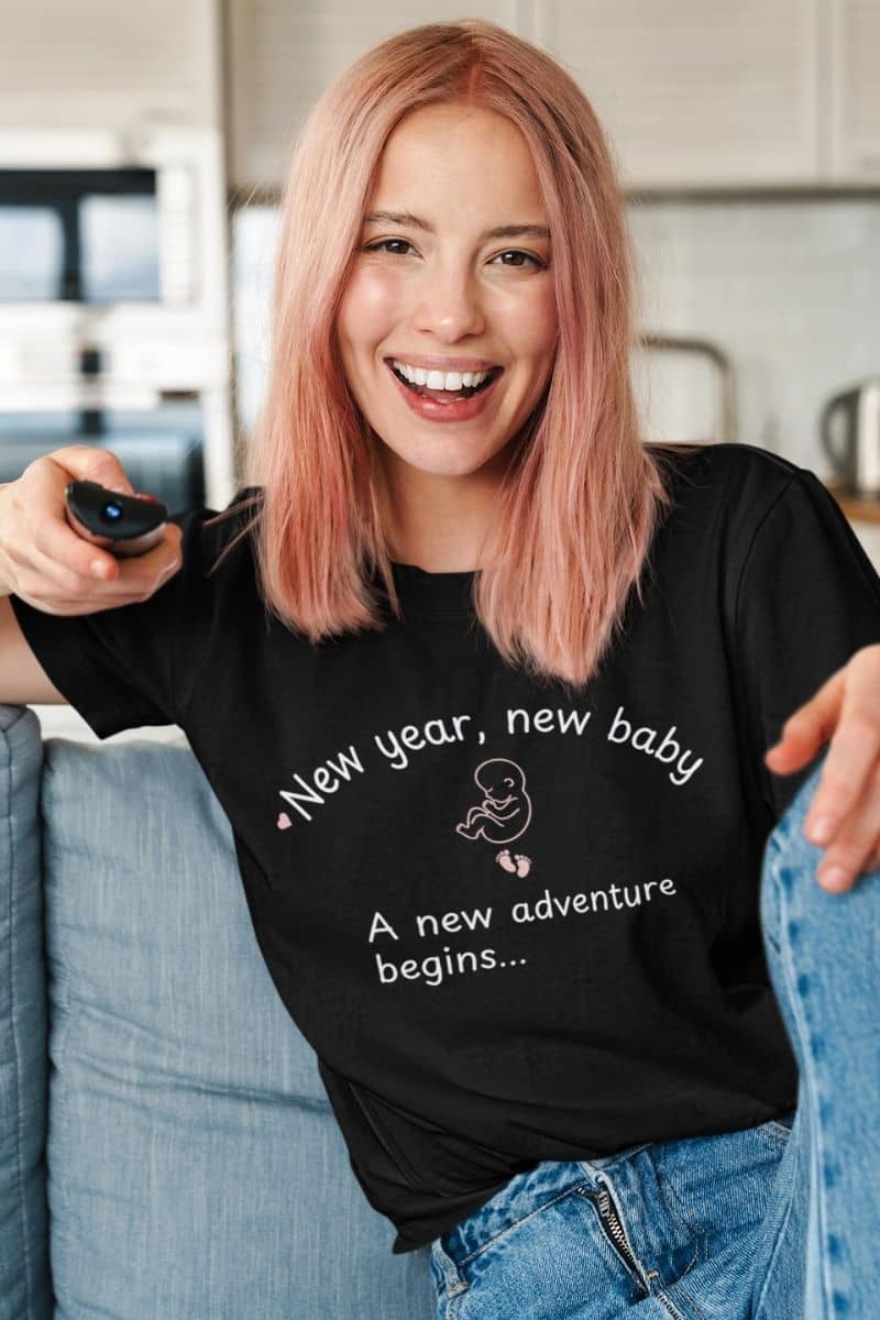New parents shirt with a woman sitting on the sofa, wearing a black t-shirt featuring the text 'New year, new baby. A new adventure begins...'