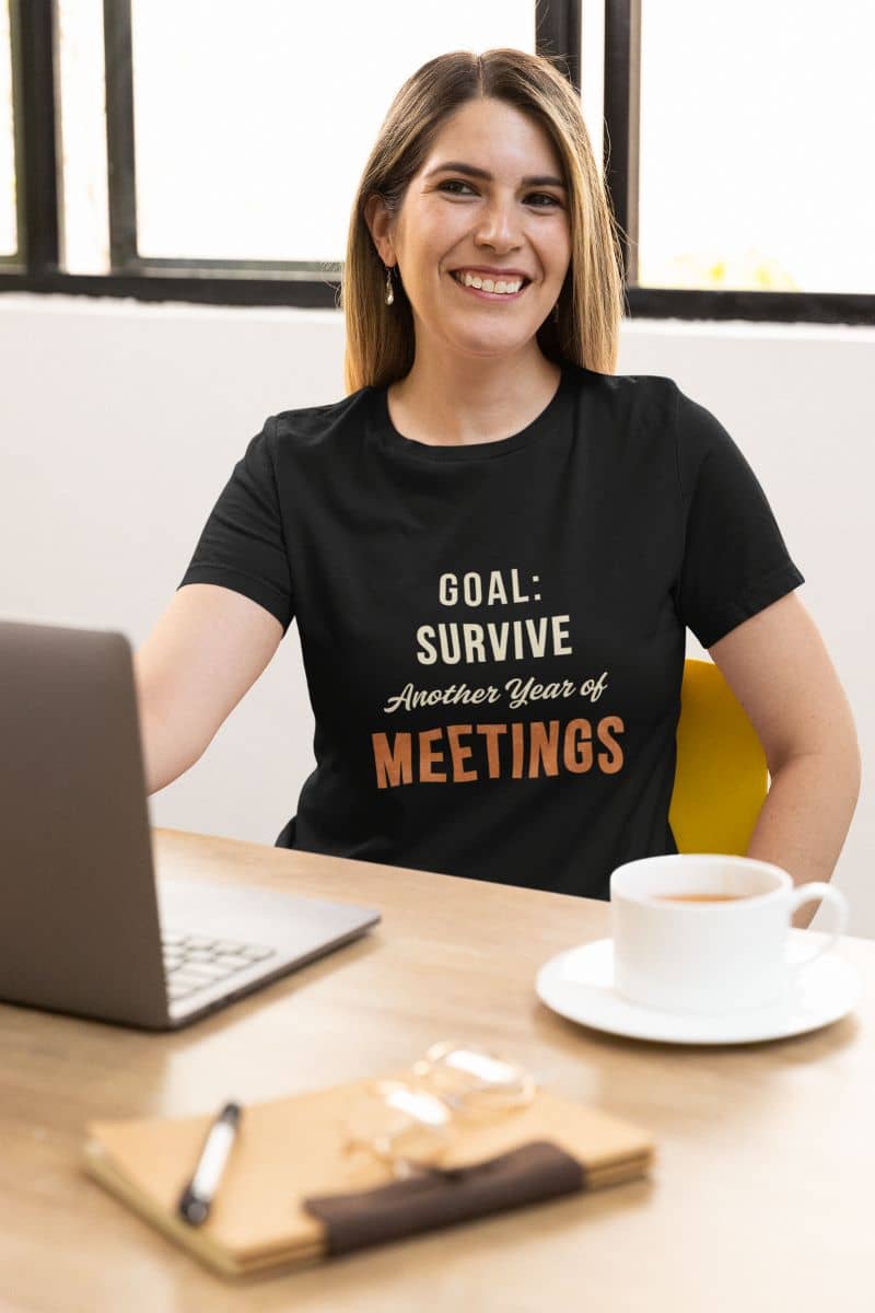 Office humor shirt with a woman wearing a black t-shirt with humorous text 'Goal: Survive Another Year of Meetings', sitting at a desk with a laptop and coffee.