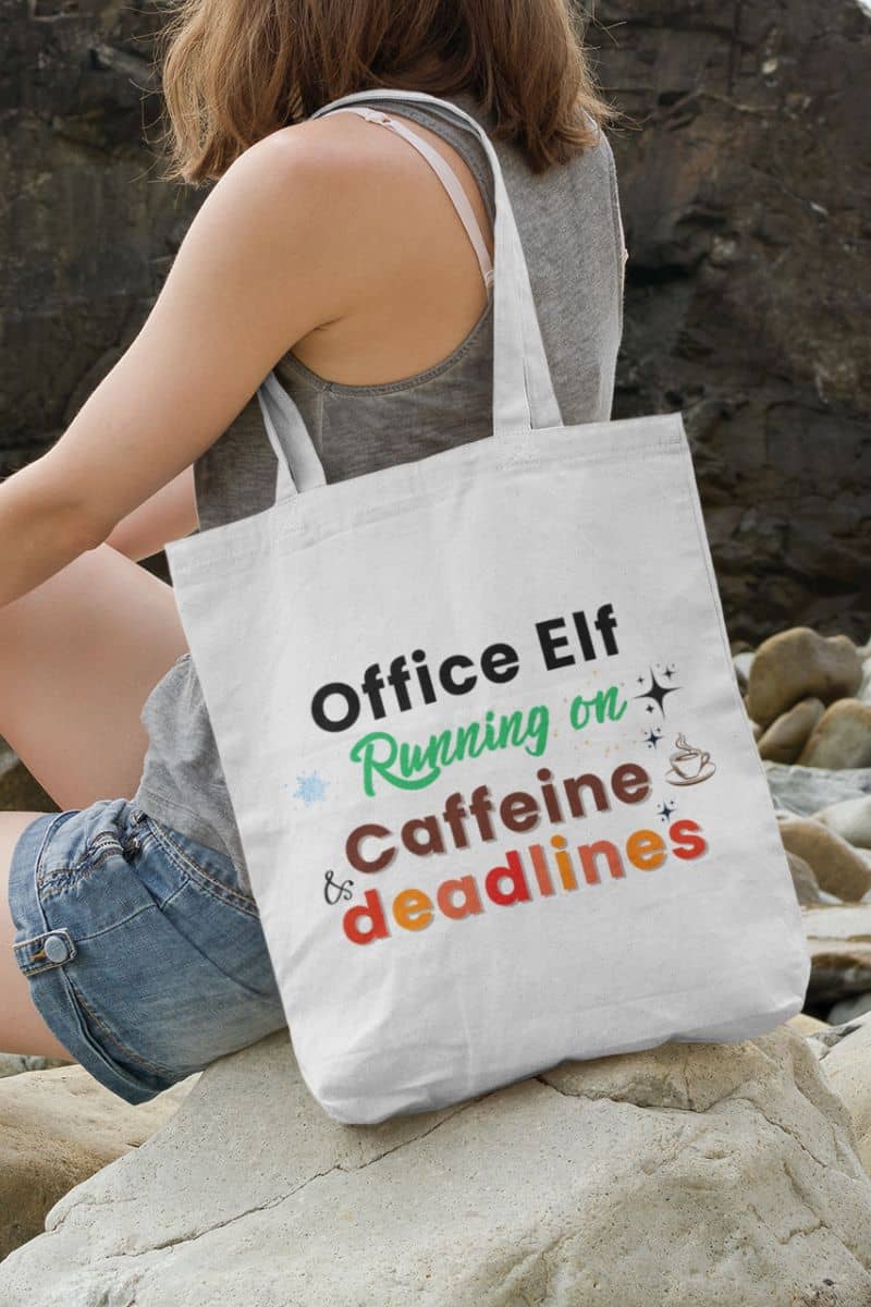 Office tote bag with a person holding a tote bag with humorous text 'Office Elf, Running on Caffeine & Deadlines' on a rocky surface